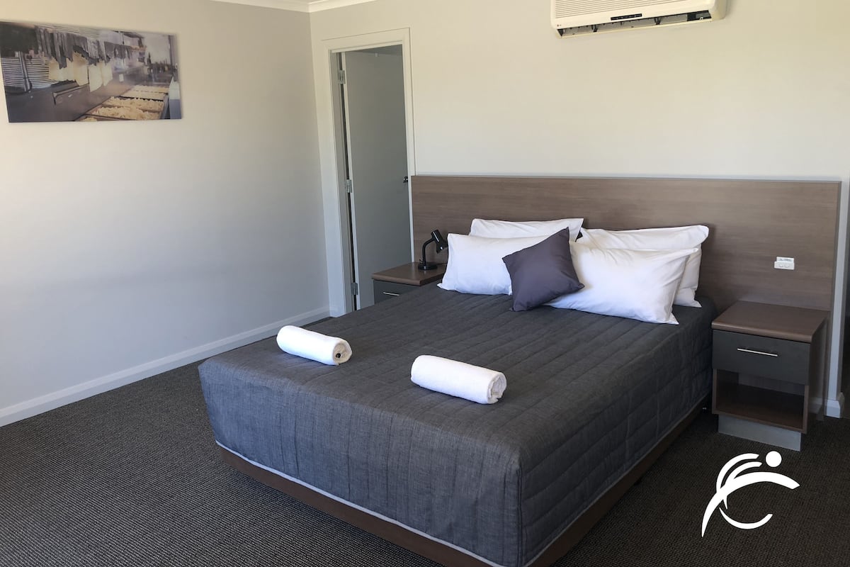 A neatly made bed with a gray quilt, two white pillows, and a rolled white towel. There is a small bedside table with a lamp, a wall-mounted air conditioner, and a door to the left.