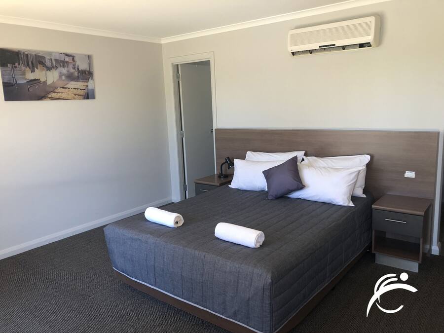 A neatly made bed with a gray quilt, two white pillows, and a rolled white towel. There is a small bedside table with a lamp, a wall-mounted air conditioner, and a door to the left.