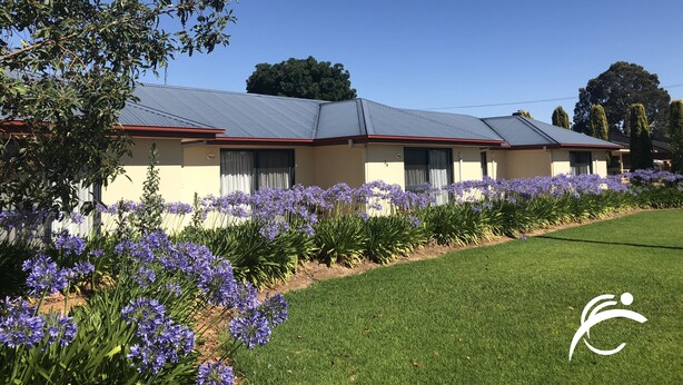 A two-story building with a flat roof, a beige facade, and a red trim. There are purple flowers in the foreground and a green lawn. - Coro Club Motel (Griffith)