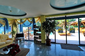 A modern interior with a sofa, a coffee table with flowers, a bookshelf, and a large window with a view of an outdoor pool area. - Bora Bora (Nessebar)
