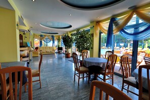 A dining area with round tables and chairs, a sofa, and a large window with a view of the outside. - Bora Bora (Nessebar)
