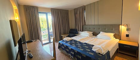 A hotel room with a bed, a desk with a chair, a small table, and a balcony with curtains.