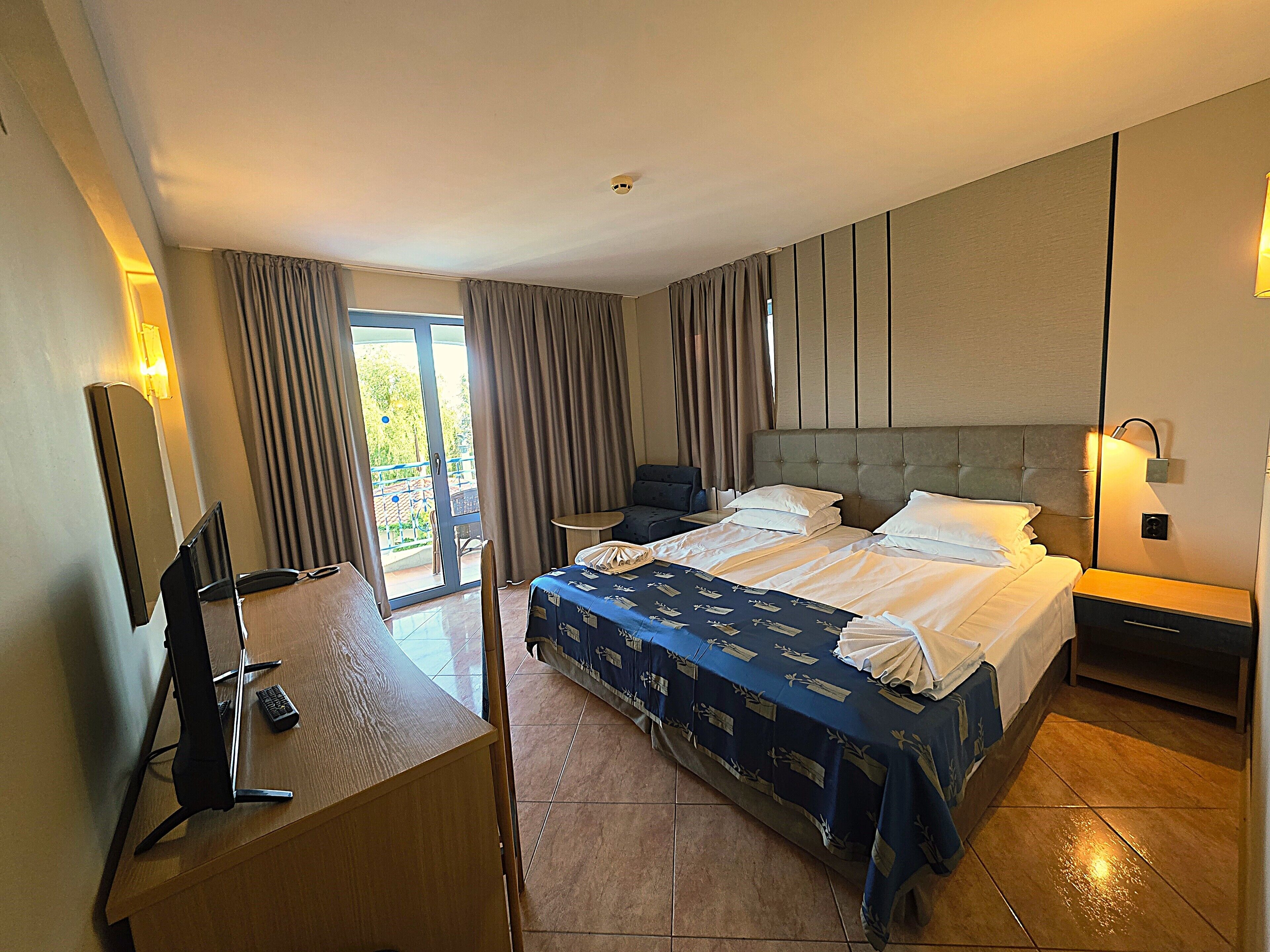 Een hotelkamer met een groot bed, een bureau met een stoel, een tafeltje en een balkon met gordijnen.