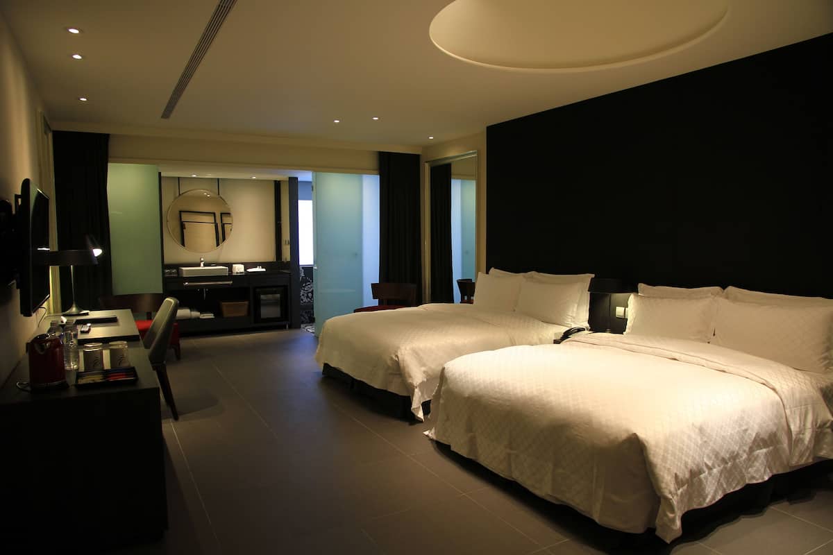 Ein Hotelzimmer mit zwei Betten, einem Schreibtisch, einem Sessel, einem Fernseher und einem Badezimmer mit Spiegel und Waschbecken.