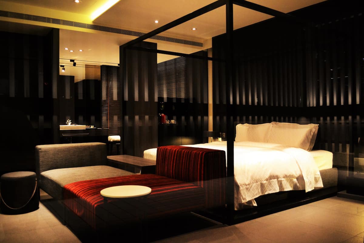 Ein modernes Hotelzimmer mit einem großen Bett, einer Sitzecke mit einem rot-schwarz gestreiften Sofa, einem kleinen runden Tisch und einer separaten Sitzecke mit einer Bank.