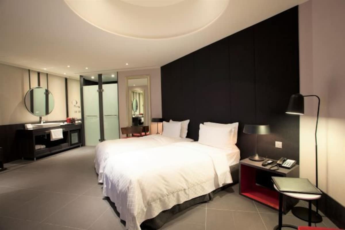 A modern hotel room with a large bed, a bedside table with a lamp, a desk with a phone, and a bathroom with a mirror and sink.