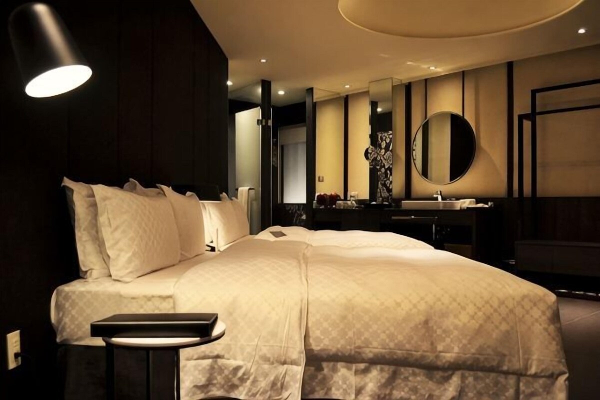 A modern hotel room with a large bed, a bedside table, a mirror, and a bathroom with a sink and mirror.