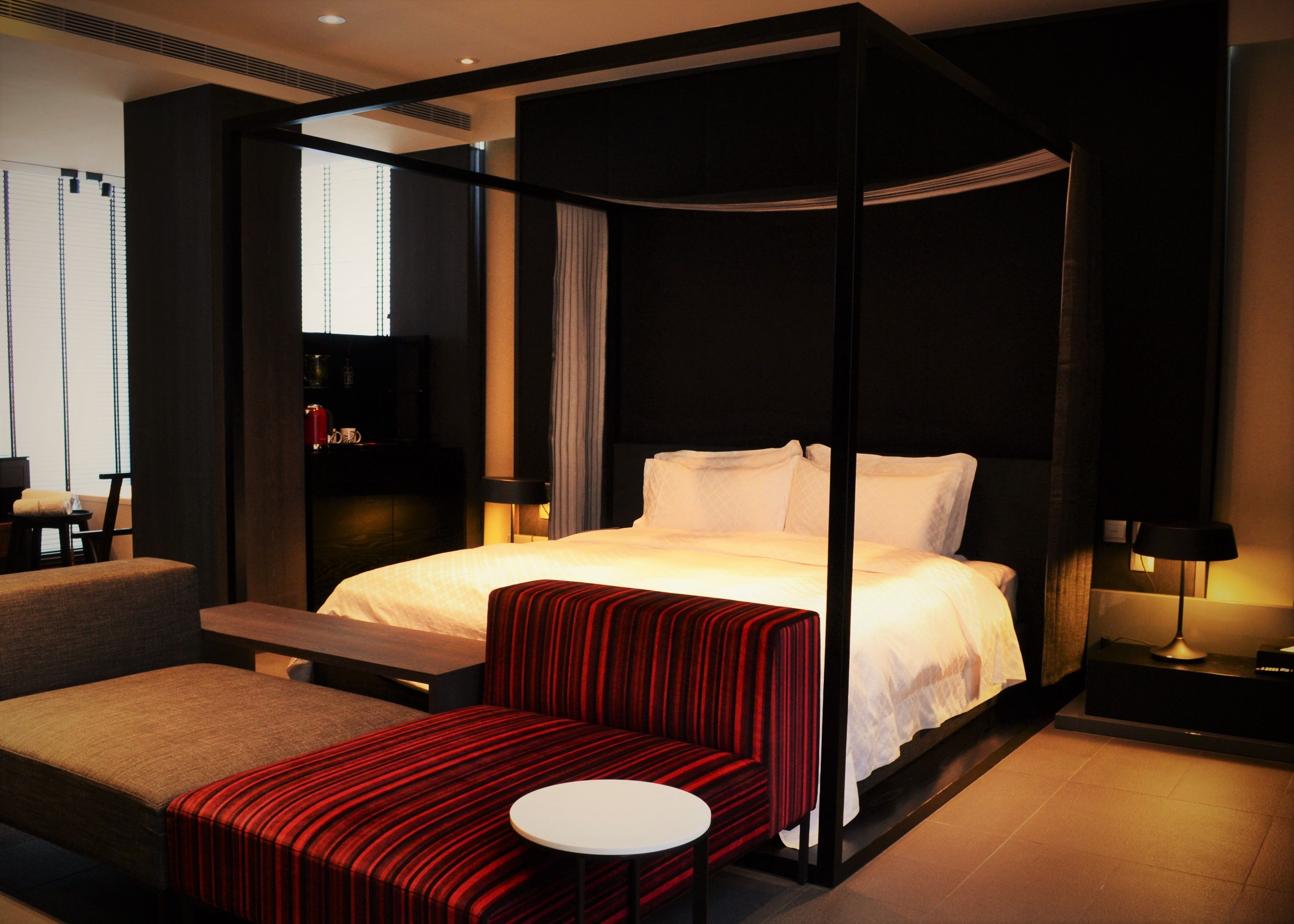 Una habitación de hotel moderna con una cama con dosel, un banco tapizado de rojo, una pequeña mesa redonda y un televisor sobre un soporte.