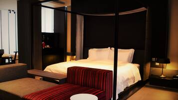 Una habitación de hotel moderna con una cama con dosel, un banco tapizado de rojo, una pequeña mesa redonda y un televisor sobre un soporte.