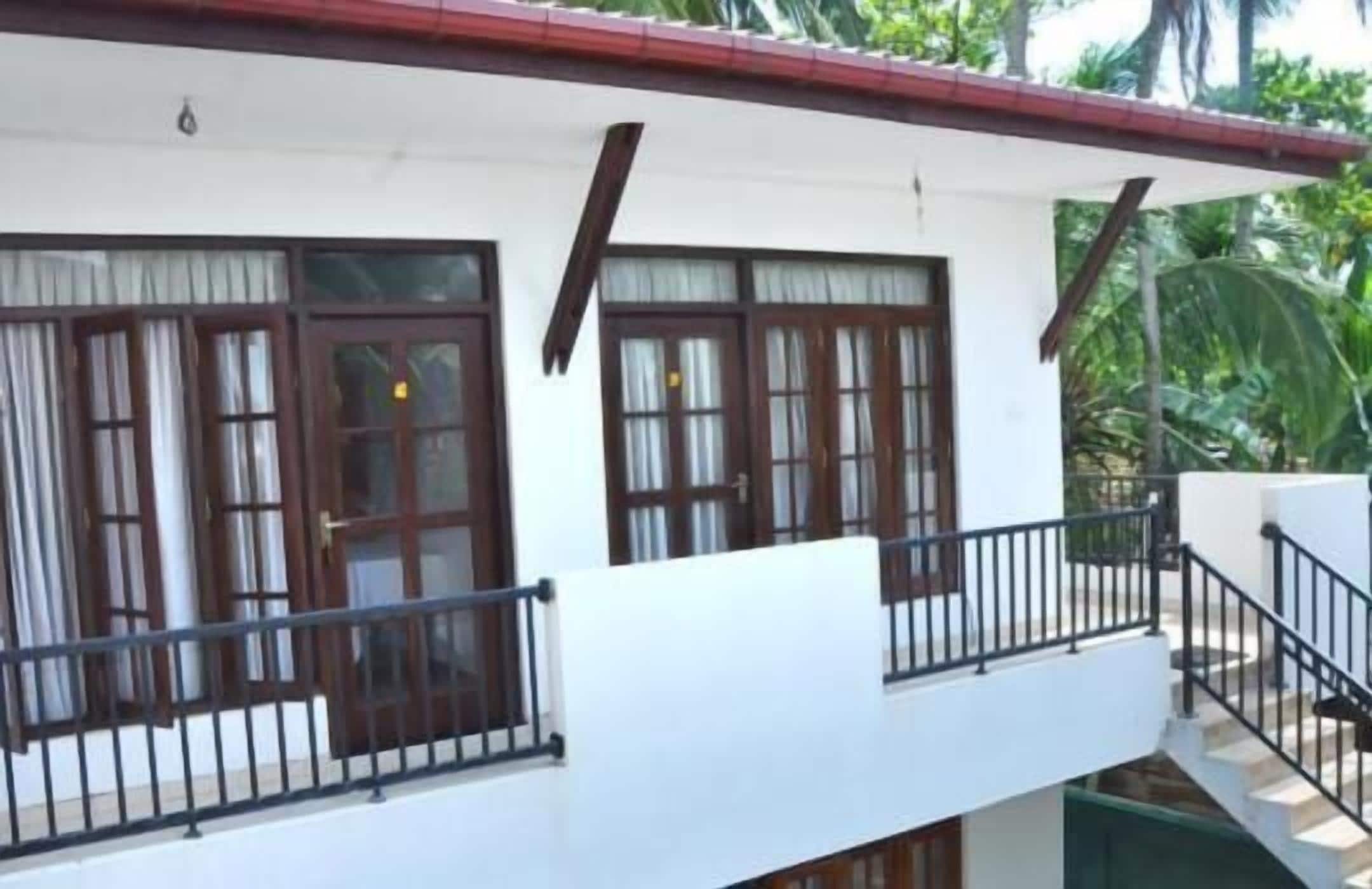 Un bâtiment à deux étages, à l’extérieur blanc et au toit rouge. Il est doté d’un balcon avec des balustrades noires et de nombreuses fenêtres avec des cadres en bois et des rideaux.