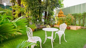 A garden with white metal chairs and a table, surrounded by lush greenery and a small decorative structure.