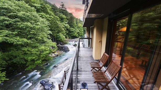 Ein Balkon mit Blick auf einen Fluss und einen Wald, mit Holzstühlen und einer Glasreling.