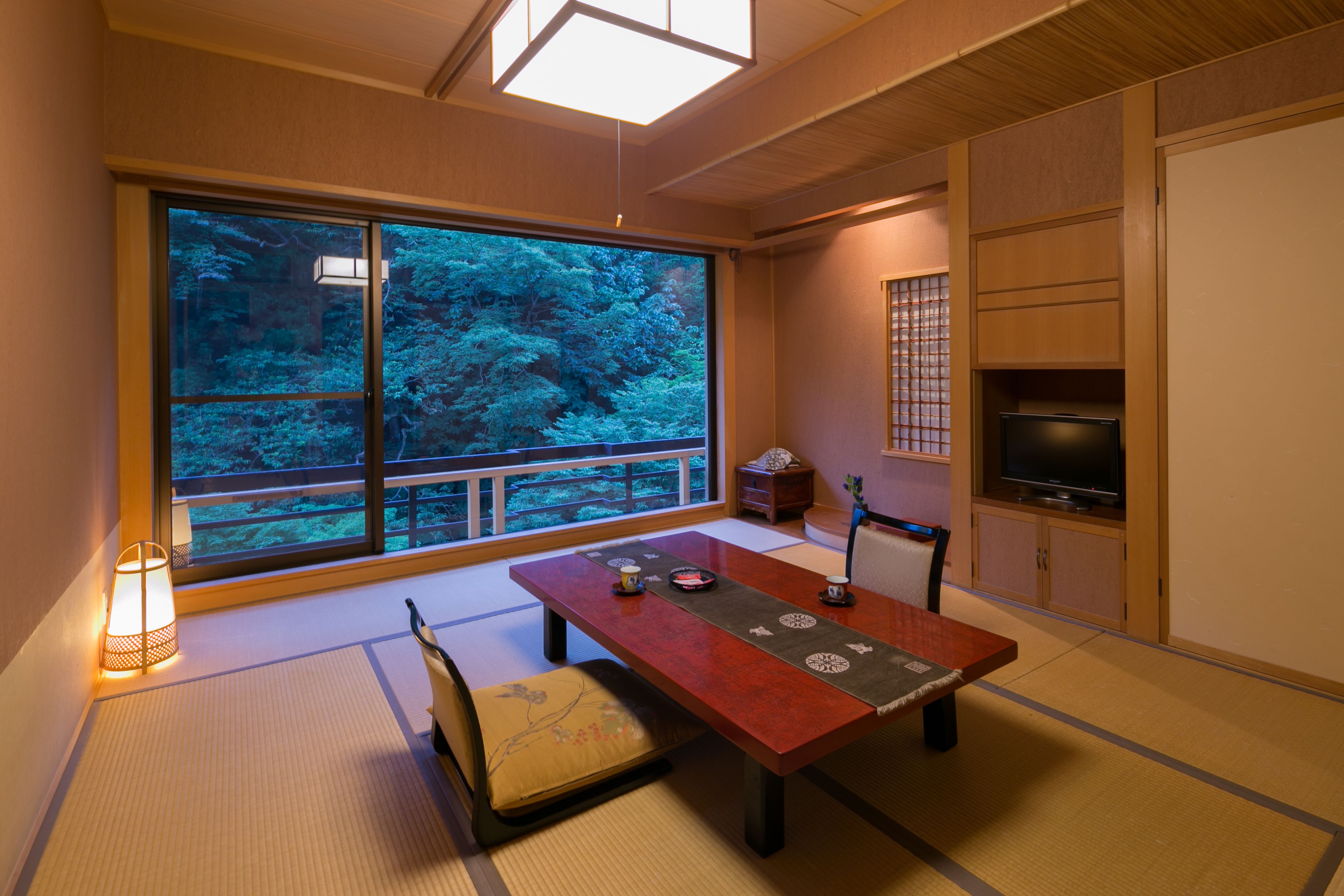 A traditional Japanese room with tatami flooring, a low wooden table, a television, and a large window with a view of trees.