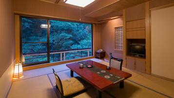 A traditional Japanese room with tatami flooring, a low wooden table, a television, and a large window with a view of trees.