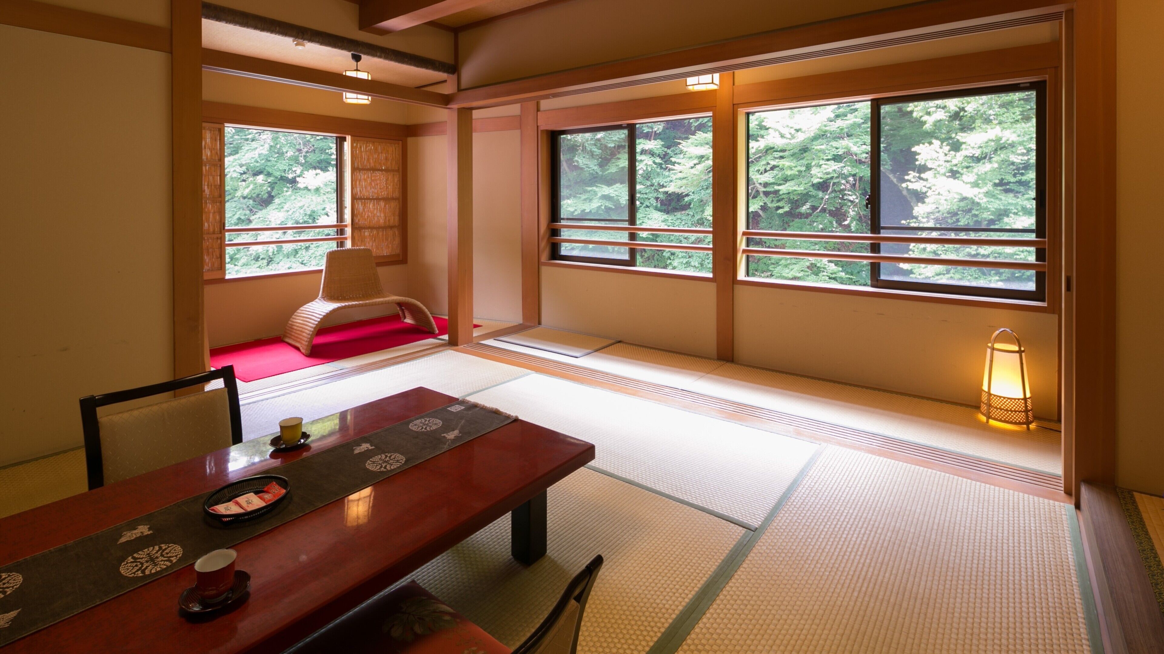 A traditional Japanese room with tatami flooring, a low table with a tray, a chair, and a view of greenery through large windows.