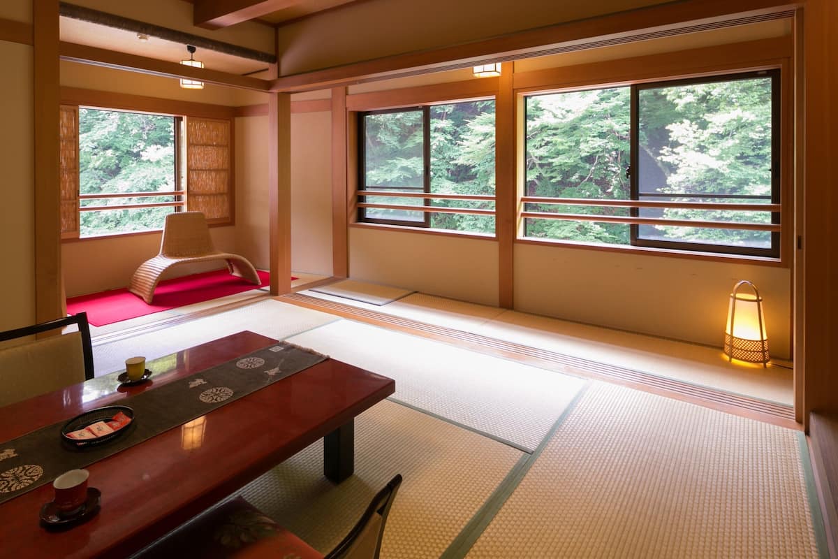A traditional Japanese room with tatami flooring, a low table with a tray, a chair, and a view of greenery through large windows.