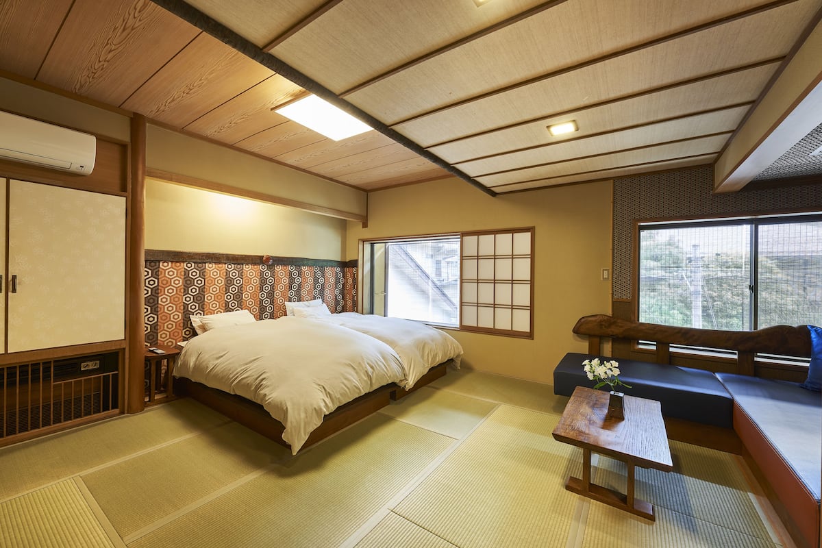A traditional Japanese room with tatami flooring, a bed with a wooden headboard, a low table with a vase, and a window with a view of trees.