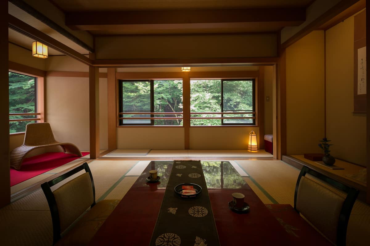 A traditional Japanese room with tatami flooring, a low table with a tray, and a view of a forest through large windows.