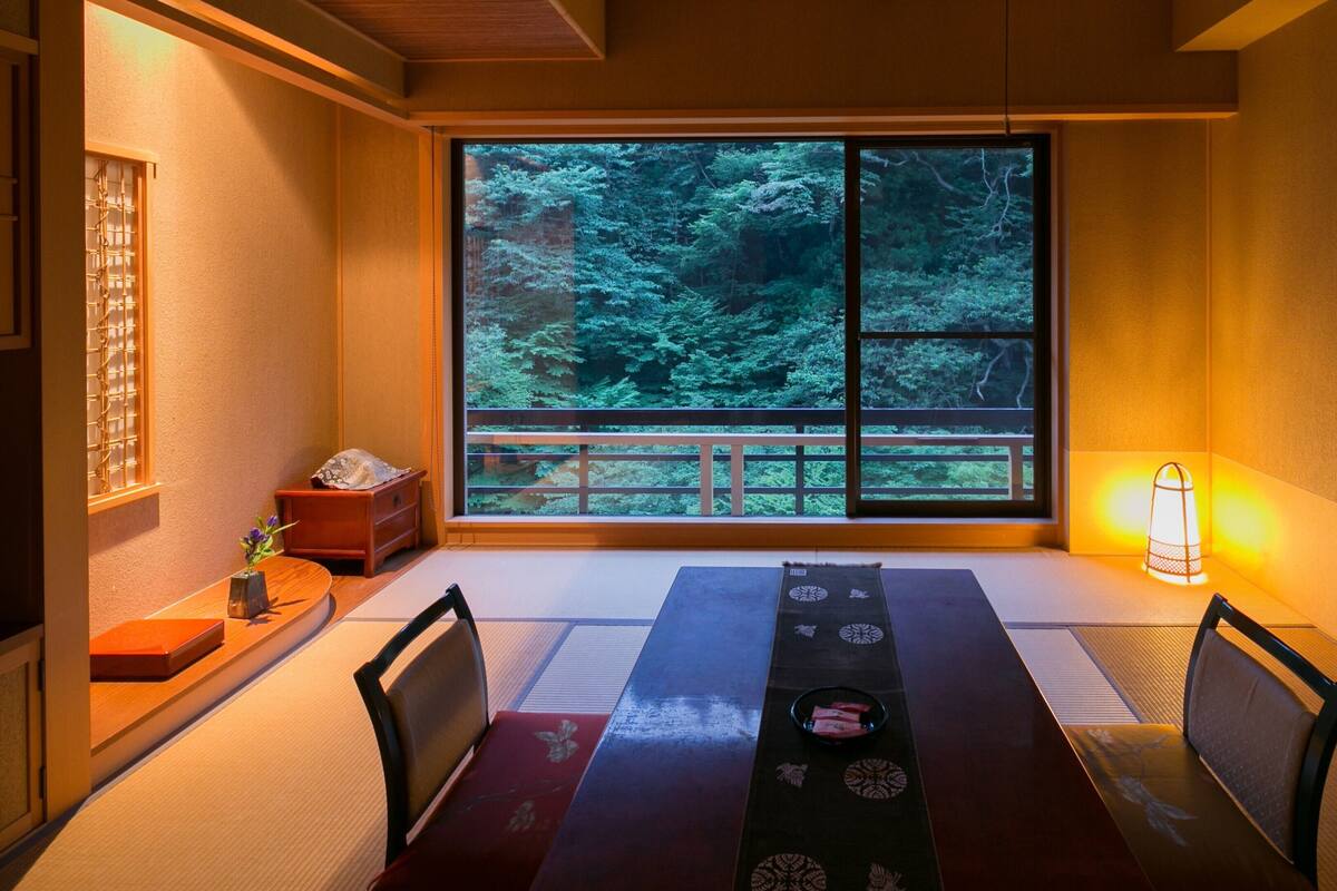 Una habitación tradicional japonesa con suelo de tatami, una mesita baja con una bandeja, sillas, una ventana con vistas a un bosque y un farol.