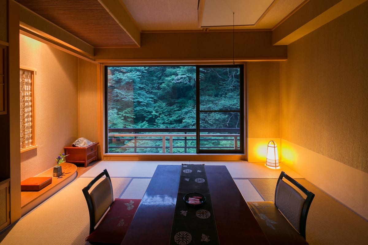 A traditional Japanese room with tatami flooring, a low table with a placemat, and a large window overlooking a forest.