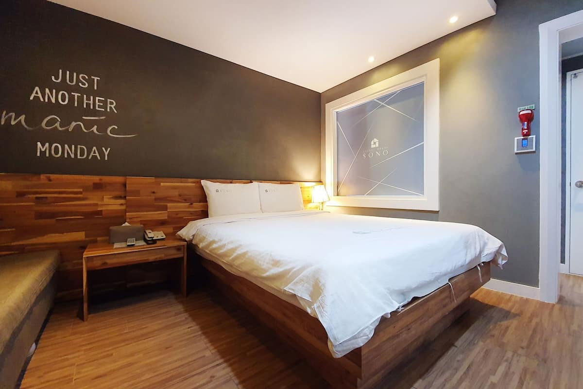 Ein modernes Hotelzimmer mit einem Holzkopfteil, einem Bett mit weißen Bettwäsche, einem Nachttisch mit Lampe und einer Wand mit motivierendem Text.