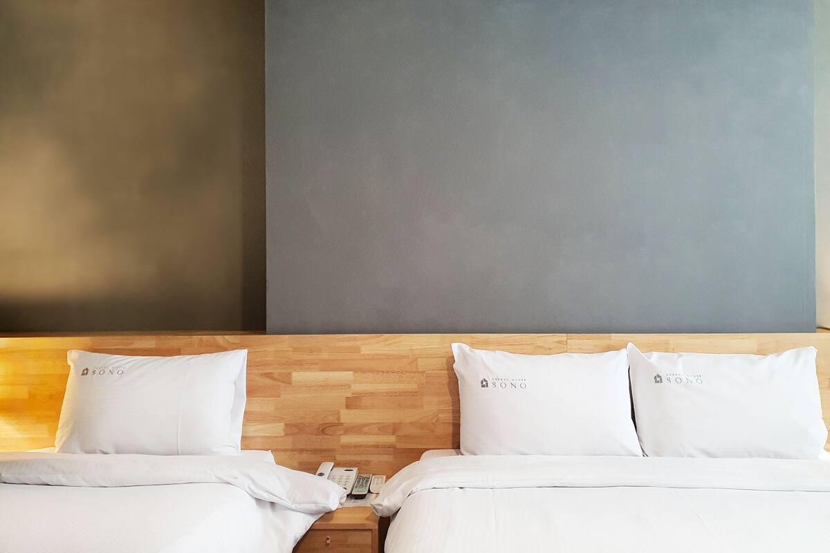 Ein Hotelzimmer mit zwei Betten, jeweils mit einem weißen Kopfkissen und einer Decke. Es gibt ein hölzernes Kopfteil, einen Nachttisch mit einem Telefon und eine Wand mit einer grauen Verkleidung.