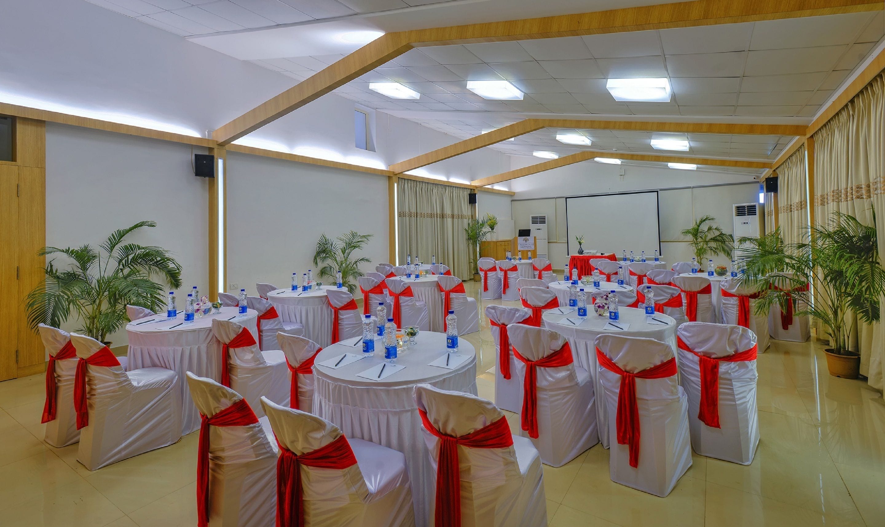 A conference room with multiple round tables, each with white tablecloths and red sashes, surrounded by chairs with white covers and red sashes.