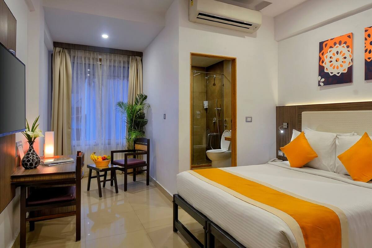 A hotel room with a bed, a desk with a chair, a small table with a vase and fruit bowl, a shower area, and two wall art pieces.