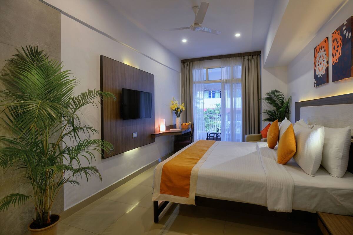 A hotel room with a bed, a flat-screen TV, a ceiling fan, a desk with a lamp, and a large window with curtains.