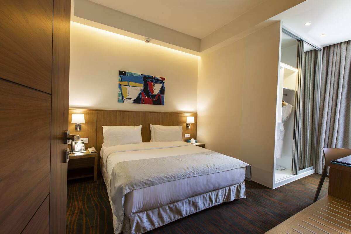 A hotel room with a large bed, bedside lamps, a painting on the wall, and a view of the bathroom through a glass door.