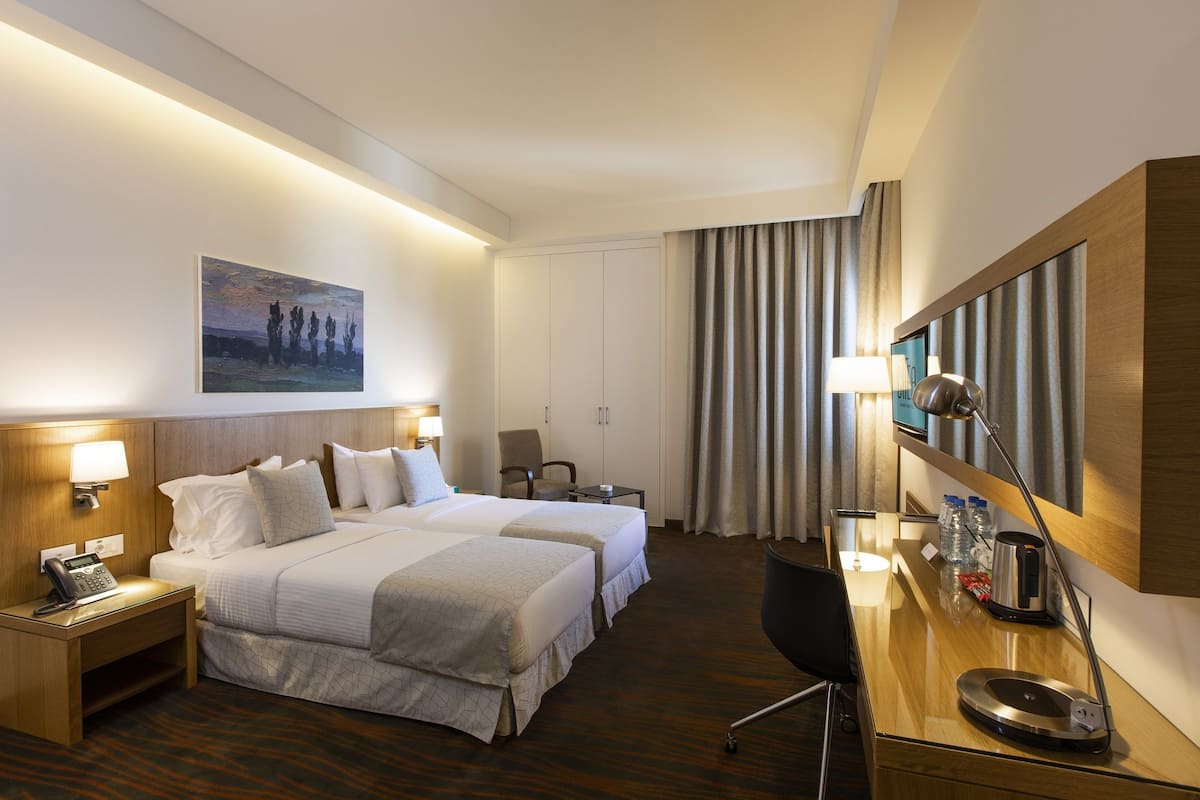 A hotel room with two beds, a desk, a chair, a telephone, a lamp, and a painting on the wall.