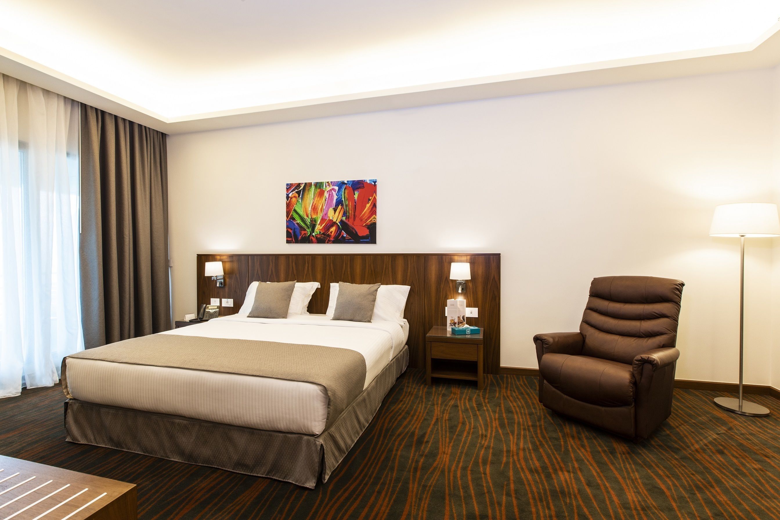 A hotel room with a large bed, a brown armchair, a nightstand with a lamp, and a colorful painting on the wall.