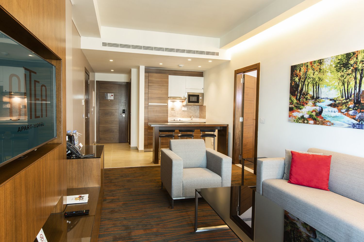 A modern hotel room with a sofa, a TV, a dining area with chairs, and a kitchen.