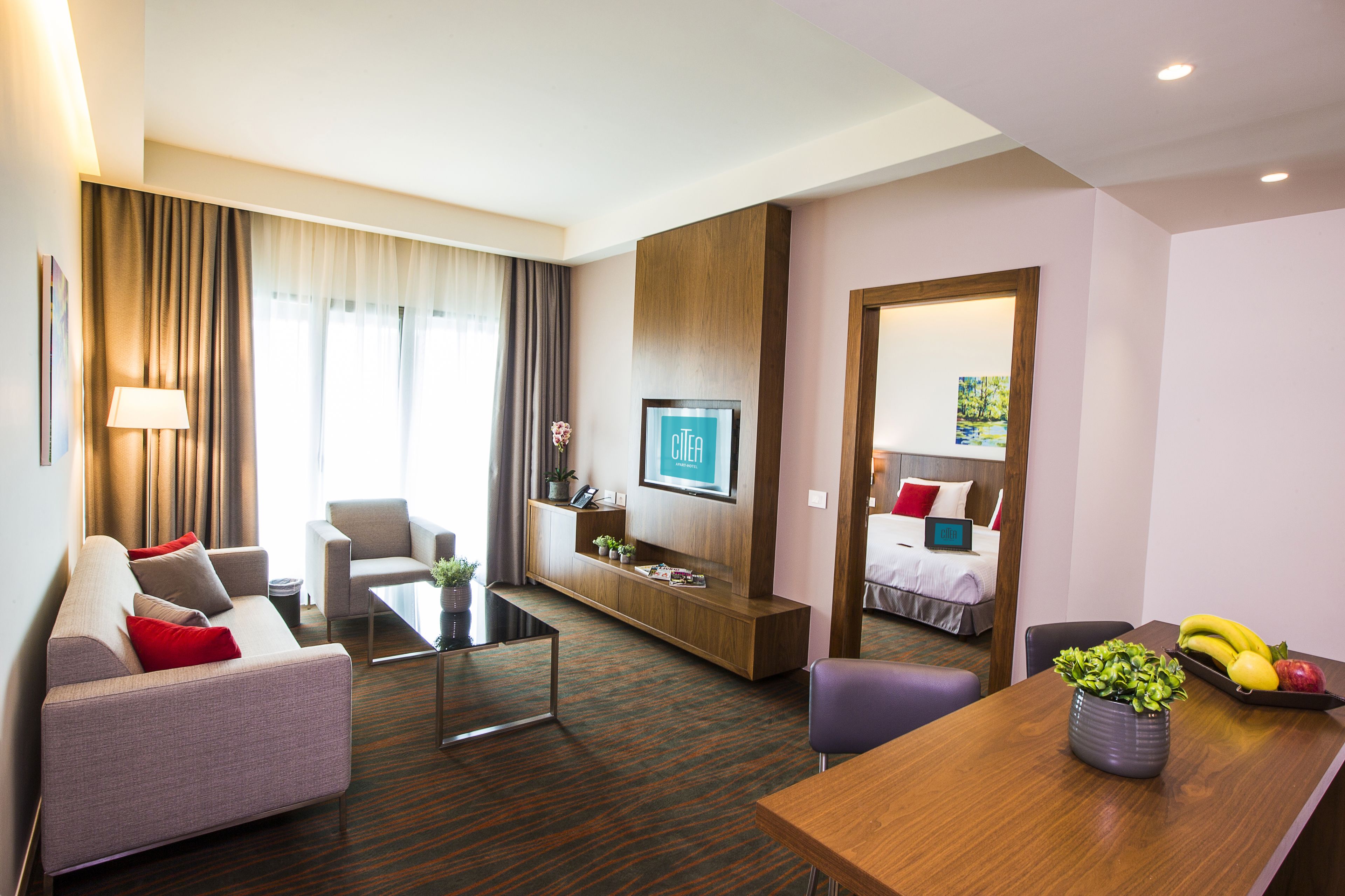 A hotel room with a sofa, armchair, coffee table, TV, and a dining area with a tray of fruit.