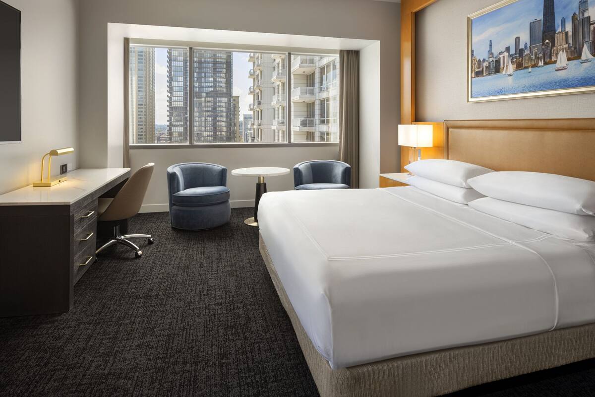 A hotel room with a large bed, two armchairs, a desk with a lamp, a small round table, and a window with a city view.
