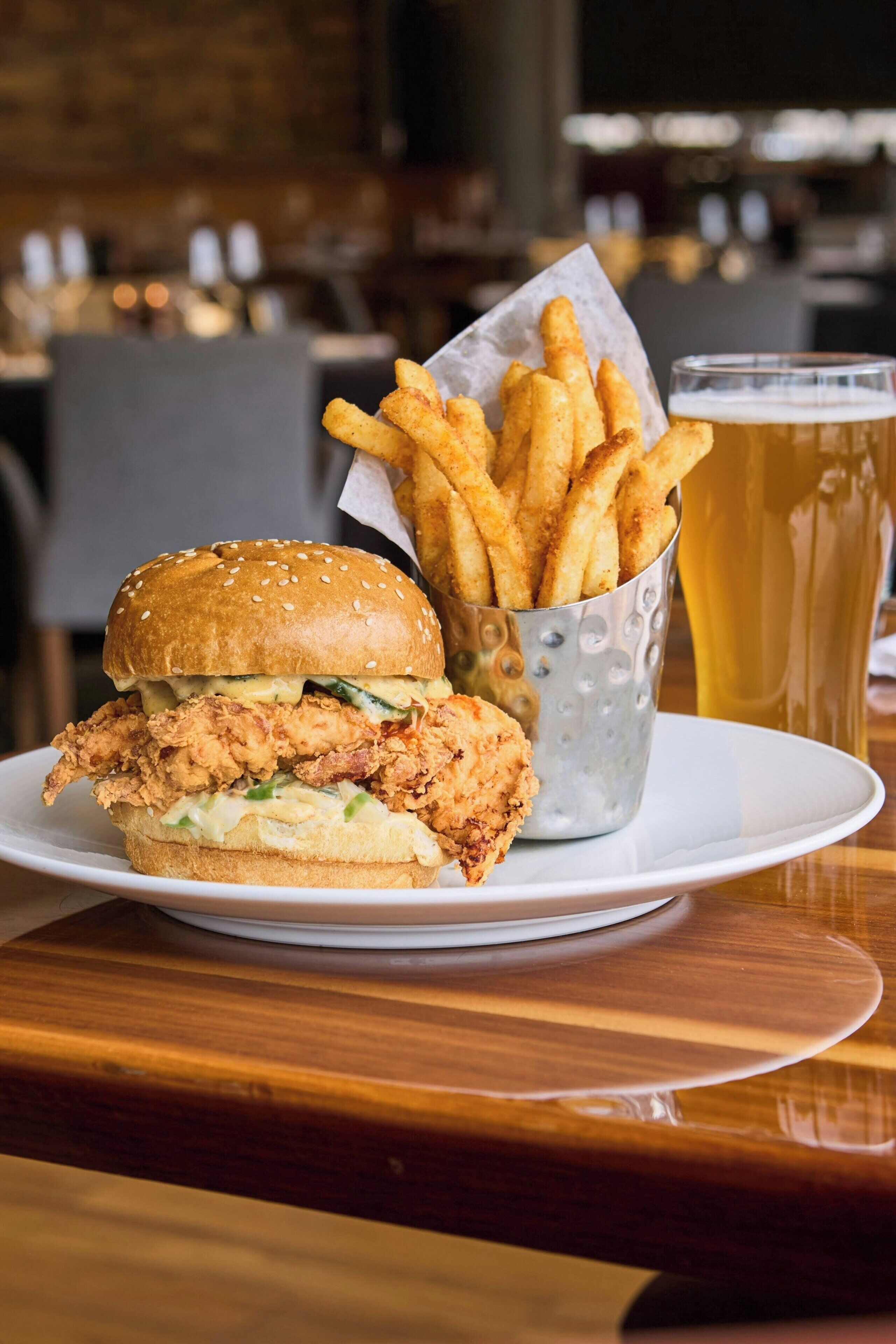 A plate with a fried chicken sandwich and a basket of fries, accompanied by a glass of beer on a wooden table.