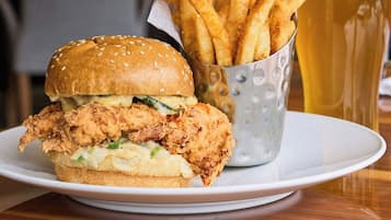 A plate with a fried chicken sandwich and a basket of fries, accompanied by a glass of beer on a wooden table.