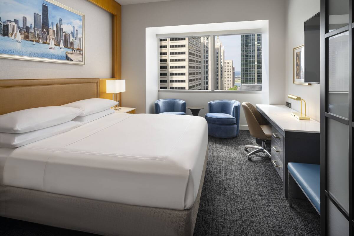 A hotel room with a large bed, a desk with two chairs, a window with a city view, and a framed picture on the wall.