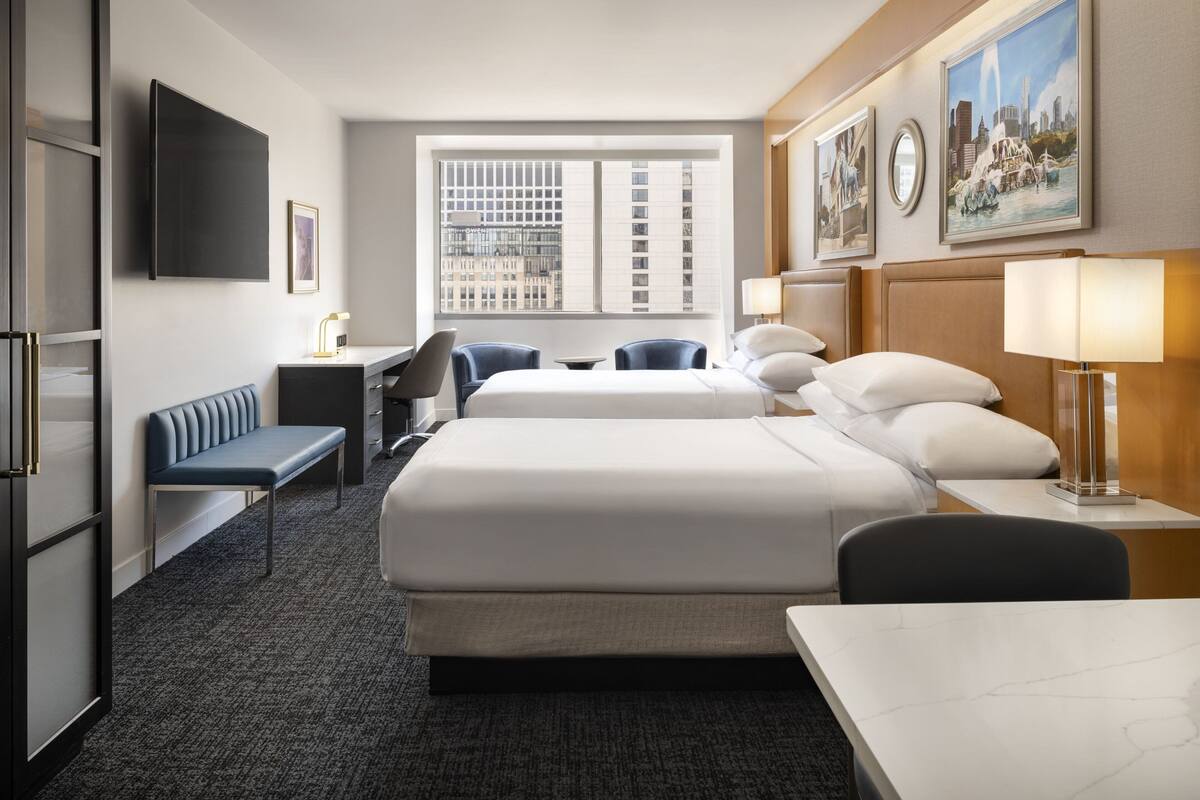 A hotel room with two beds, a desk, a bench, a TV, and a window with a city view.