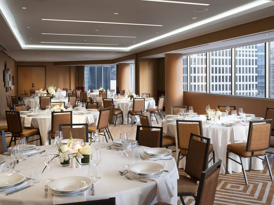 Un comedor de hotel con varias mesas dispuestas para comer, amplias ventanas que ofrecen vista a la ciudad y un diseño de techo moderno.