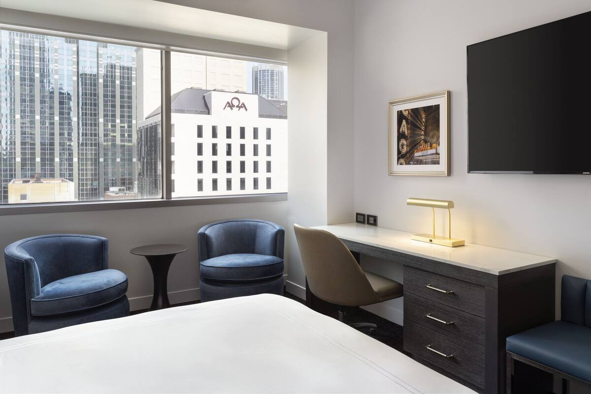 A hotel room with a bed, two blue armchairs, a desk with a lamp, a TV, and a window with a city view.