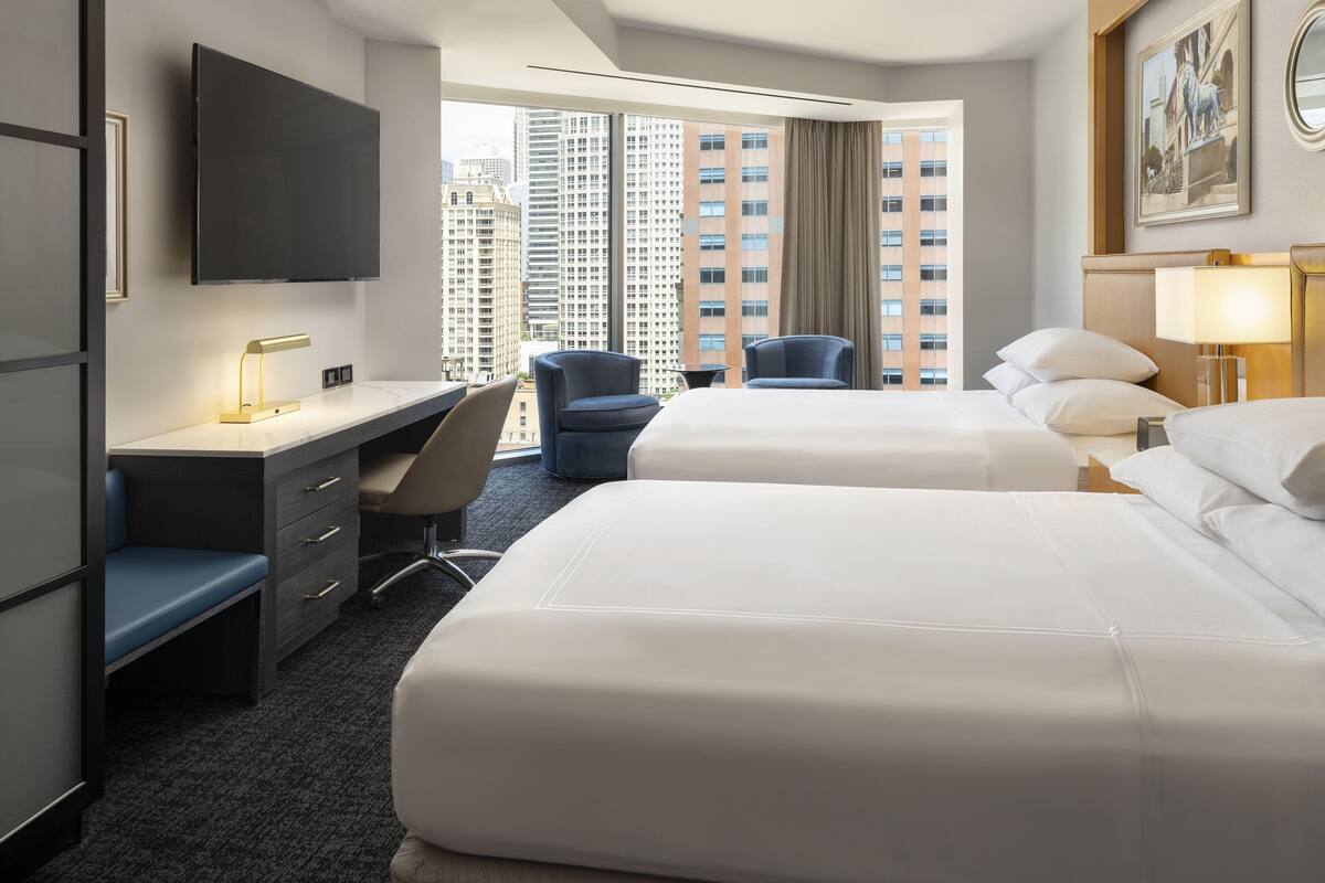 A hotel room with two beds, a desk, a chair, a TV, and a large window with a city view.