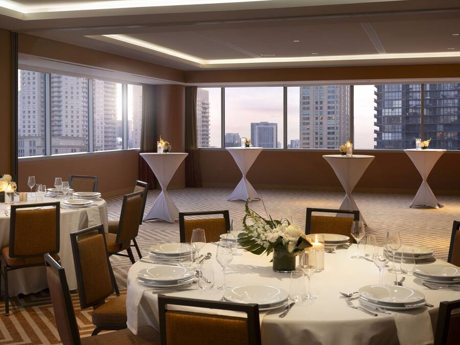 A banquet hall with round tables set for dining, large windows with a cityscape view, and a carpeted floor.
