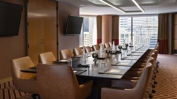 A conference room with a long table, multiple chairs, a flat-screen TV, and large windows with a city view.