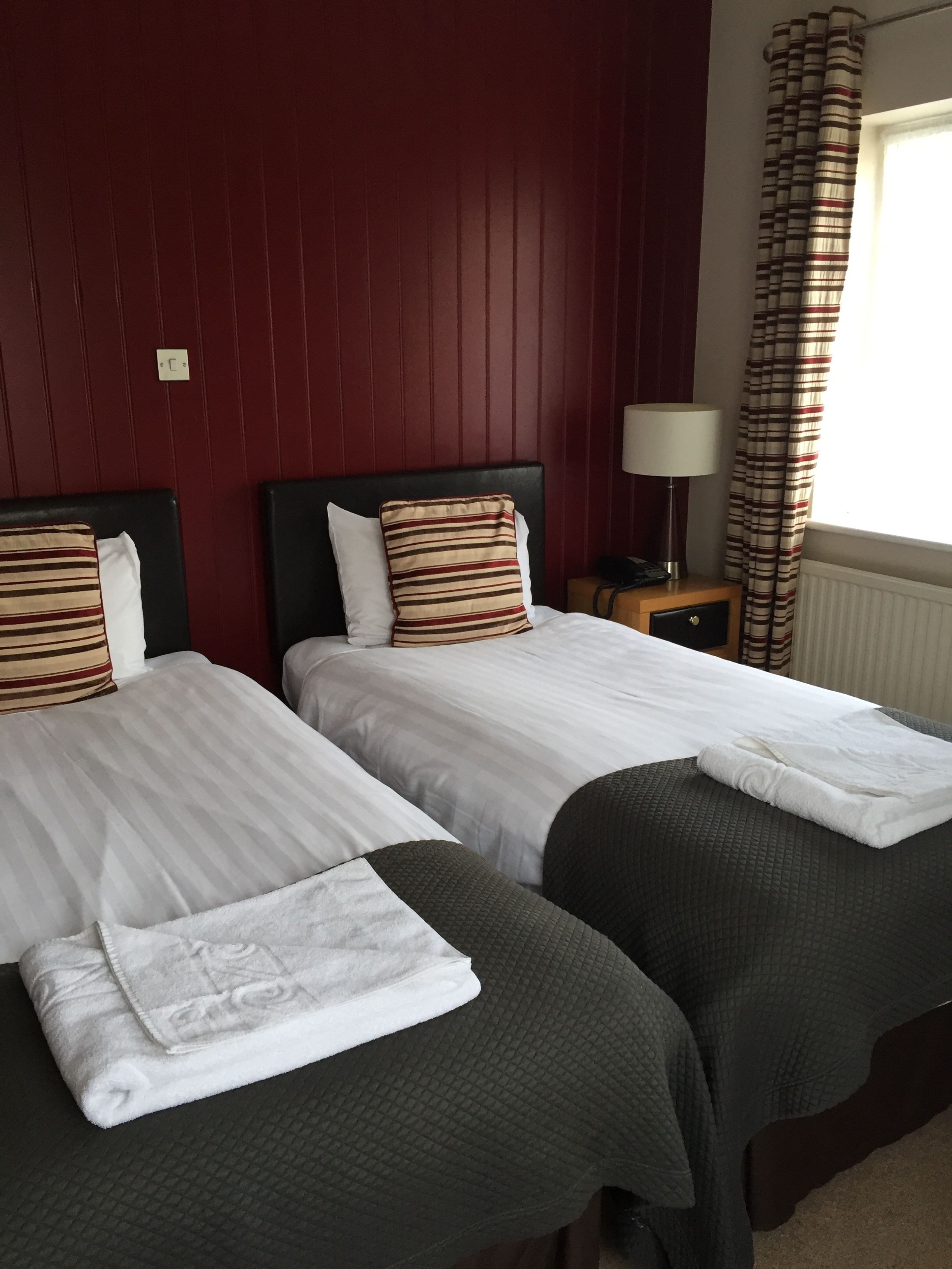 A hotel room with two beds, each with white linens and striped pillows. There is a nightstand with a lamp and a telephone. The room has a red wall and a window with striped curtains.