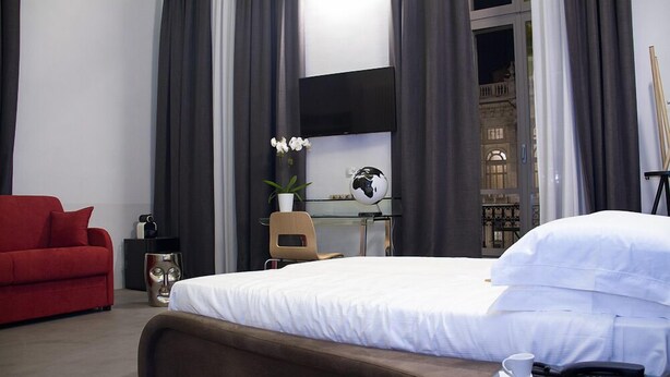 A modern hotel room with a large bed, a red sofa, a glass table with a globe and flowers, and a view of a building through the window. - Piazza Castello Suite (Turin)