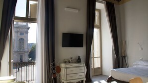 A hotel room with a bed, a dresser, a TV, and a view of a historic building through large windows. - Piazza Castello Suite (Turin)