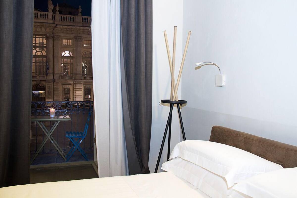 Une chambre d’hôtel avec un lit, une table de chevet avec une lampe, une vue sur un immeuble par la fenêtre et un balcon avec une table et des chaises.