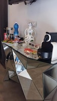 A dining table with a glass top and geometric metal base, surrounded by various food items and a coffee machine.