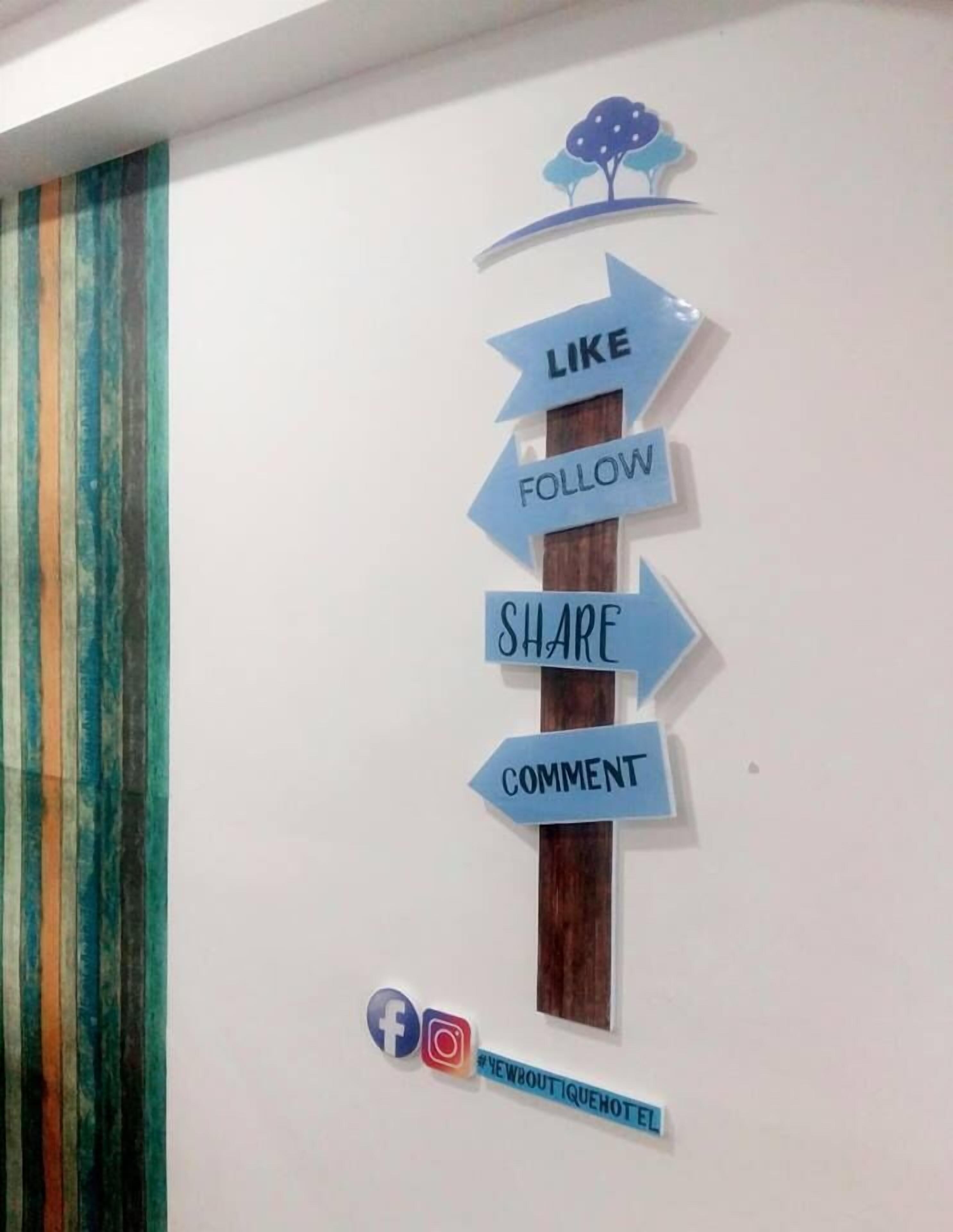 Una pared con un letrero vertical que muestra flechas indicando las palabras 'LIKE', 'FOLLOW', 'SHARE' y 'COMMENT'. Debajo del letrero, hay iconos de redes sociales para Facebook e Instagram, y un hashtag '#YEWABOUTQUEHOTEL'.